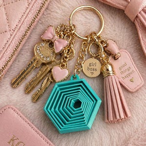 Puede incluir: Un llavero dorado con múltiples dijes, que incluyen un lazo rosa, un corazón y una forma geométrica turquesa. El llavero también presenta una borla rosa y una etiqueta que dice "good things happen".