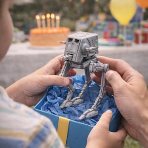 May include: A gray AT-ST model from Star Wars is presented in a blue gift box with blue tissue paper. The toy is held by a person at a birthday party with a cake and balloons in the background.