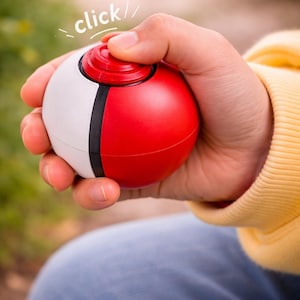 Puede incluir: Un juguete esférico rojo y blanco con una franja negra, sostenido en una mano. La parte superior del juguete es roja con un botón circular. La palabra "click" está escrita encima del juguete.