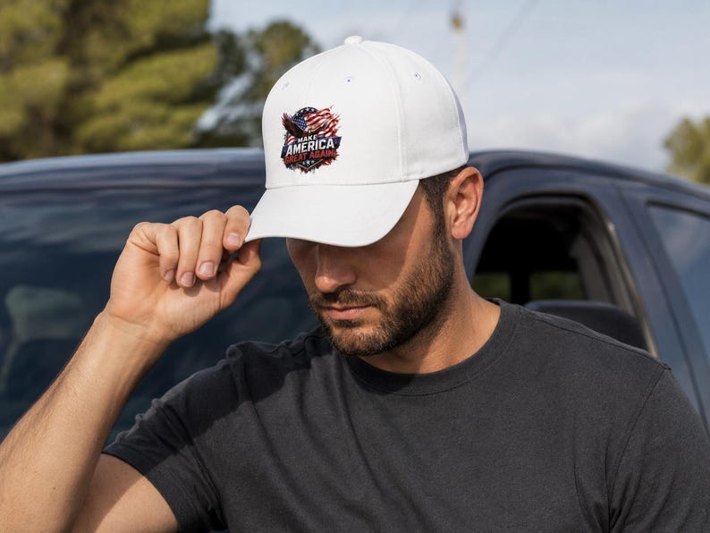May include: White baseball cap with a curved brim. The cap features a graphic of an eagle, American flag, and the text "MAKE AMERICA GREAT AGAIN" in red, white, and blue. The cap is being held by a person.