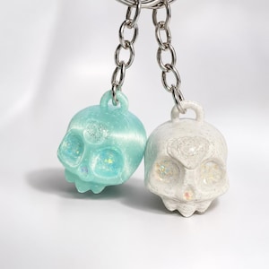 Puede incluir: Dos llaveros con forma de calavera unidos a anillas y cadenas de metal plateado. Una calavera es de color verde menta translúcido con purpurina iridiscente en las cuencas de los ojos y la frente. La otra es blanca opaca con textura moteada.
