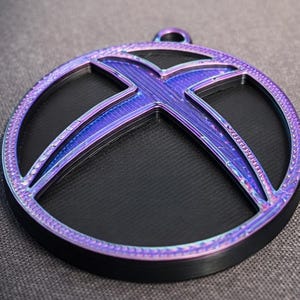 Puede incluir: Colgante redondo negro y morado con un marco circular y un diseño estilizado de 'X' en el centro. El colgante tiene una superficie texturizada y un pequeño lazo para colgar. El morado tiene un brillo metálico. Diámetro: aprox. 7 cm.