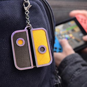 Puede incluir: Un llavero con diseño estilizado que se asemeja a un mando de videojuegos. Un lado es amarillo, el otro gris oscuro, ambos con detalles circulares. El llavero está sujeto a una mochila con una cadena y un cierre plateados. Medidas en cm.