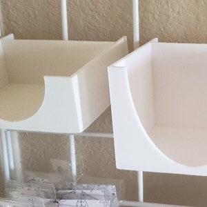 Peut inclure: Trois bacs de rangement en plastique blanc avec un bord avant incurvé. Les bacs sont montés sur un rack en fil métallique blanc.