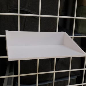 Puede incluir: Una estantería blanca en ángulo diseñada para fijarse a una rejilla metálica. La estantería está hecha de un material sólido y opaco y tiene un diseño simple y funcional. La estantería se muestra fijada a una rejilla metálica blanca.