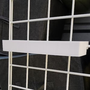 Op de afbeelding: Een witte rechthoekige plank bevestigd aan een wit draadrek. De plank is gemaakt van een solide, ondoorzichtig materiaal en is ontworpen om items vast te houden. Het rek is gemaakt van witte metalen draden en de achtergrond is een donkere grijze stof.