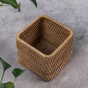 May include: A square woven wicker basket in a natural brown colour. The basket is empty and sitting on a light grey surface.