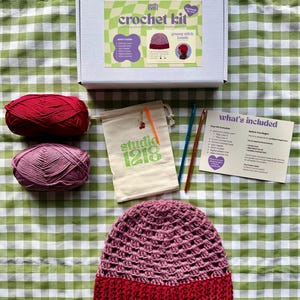Könnte beinhalten: Ein Häkelset in einer weißen Schachtel mit grün kariertem Etikett, bestehend aus zwei Garnknäueln in Rot und Rosa, einem beigen Beutel mit dem Studio 1218-Logo, zwei Häkelnadeln, einer Kunststoffnadel und einer gedruckten Anleitungskarte.