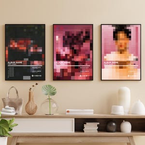 May include: Three framed art prints with pixelated images in black, red, and pink hues. Each print features text including "Album Name" and "Artist Name", along with release date and album length details. The prints are displayed above a wooden console.