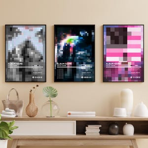 May include: Three framed art prints with pixelated designs in black and white, multicolored, and pink hues. Each print features the text "ALBUM NAME" and "ARTIST NAME" along with other details. The prints are displayed on a wooden console table.