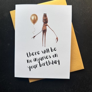 Op de afbeelding: Een witte wenskaart met de tekst "there will be no injuries on your birthday" in zwart script. De kaart toont een bruin wezen met een gouden ballon. De kaart ligt op een gouden envelop.