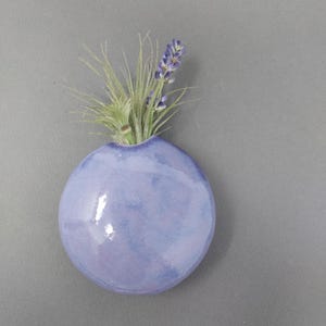 May include: A round, light purple ceramic wall vase with a glossy finish. The vase contains a small green air plant and a single sprig of purple lavender. The object is circular with a flat back and a small opening at the top for the plants.