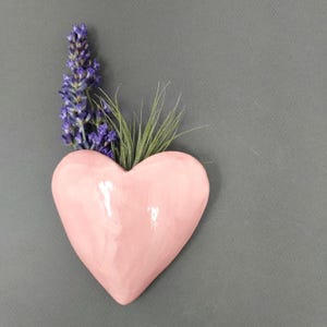 May include: A glossy, light pink heart-shaped ceramic wall planter featuring a smooth, rounded surface. The planter contains a sprig of purple lavender flowers and a small, spiky green air plant extending from the top opening of the heart.