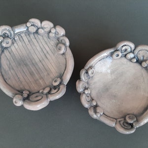 May include: Two small, ceramic dishes with a light pink glaze. The dishes have a decorative, scalloped edge with raised, rounded details. One dish has a textured, striped surface.