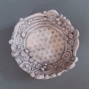 May include: A small, white ceramic bowl with a textured, dotted surface and a decorative, raised rim. The rim features swirls and bumps.