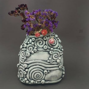 May include: A light blue ceramic vase featuring a textured design of swirls and raised circles. The vase contains dried purple and red flowers. The decorative vase is a unique home decor item, with a height of approximately 15 cm.