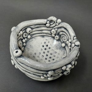 May include: A handcrafted ceramic bowl with a unique, textured design. The bowl is primarily light gray with darker gray accents, featuring a series of raised, organic shapes and a perforated interior. The rim has a wavy, flowing pattern.