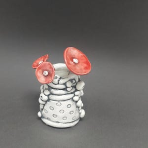 May include: A small ceramic vase with a white base and red flower accents. The vase has a textured surface with black detailing and white dots. Three red flowers with white centres are attached to the top.