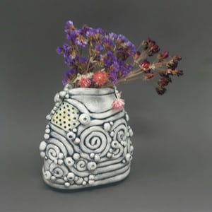 May include: A small, white ceramic vase with a textured design of spirals, dots, and raised elements. The vase holds dried purple and red flowers. The vase has a yellow dotted section. The overall design is artistic and decorative.