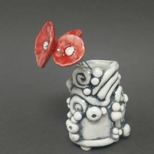 May include: A small ceramic vase with a white and grey design, holding two red poppy flowers. The vase has a textured surface with raised, circular details. The poppies have white centres.