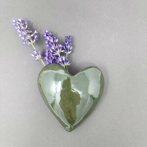 May include: A heart-shaped ceramic wall vase with a glossy olive green finish. The vase contains a small bunch of purple lavender sprigs. The object has a smooth, reflective surface and a rounded, symmetrical form, providing a simple vessel.
