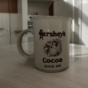 May include: A white ceramic Hershey's Cocoa mug with a brown handle and rim. The mug features the Hershey's logo, an illustration, and the words "Cocoa" and "SINCE 1895" in brown lettering. The mug is on a light-colored surface.