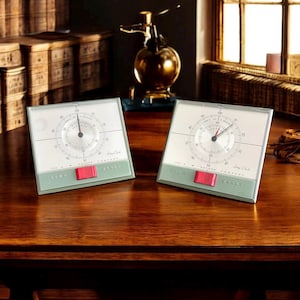May include: Two vintage-style, square clocks with a light green and red accent. The clocks have a circular gauge with numbers and hands, and the text "The A.W. Haydon Company" is printed on the bottom. The clocks are on a wooden surface.