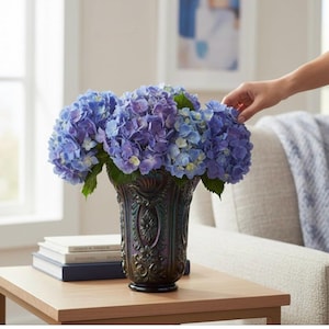 May include: A decorative vase filled with vibrant blue and purple hydrangeas. The vase has an ornate, dark-colored design and sits on a wooden table. A hand gently touches the flowers, suggesting a floral arrangement.
