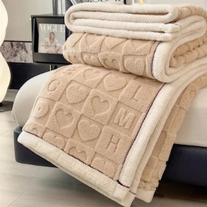 Puede incluir: Una manta suave y mullida con un borde de color crema y un centro beige con un patrón repetido de corazones y letras. La manta está doblada y se muestra en una cama.