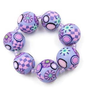 May include: A bracelet featuring round, patterned beads in shades of lavender, pink, and white. The beads display floral and checkered designs, strung together with a clear, elastic cord. The bracelet is set against a white background.