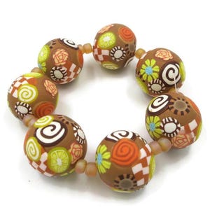 May include: A bracelet featuring brown, patterned beads. Each bead is decorated with colorful floral and geometric designs in orange, green, white, and yellow. The beads are strung together with a neutral-colored cord.