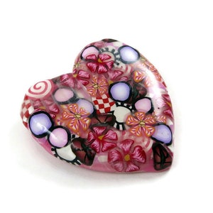 May include: A pink heart-shaped charm with a colorful floral pattern. The charm is made of polymer clay and features a variety of flowers, swirls, and polka dots.