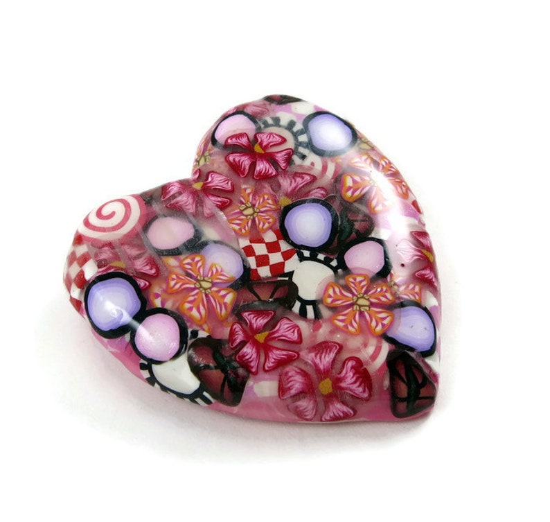 May include: A pink heart-shaped charm with a colorful floral pattern. The charm is made of polymer clay and features a variety of flowers, swirls, and polka dots.