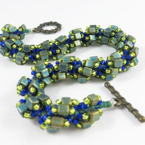 May include: A green and blue beaded bracelet with a toggle clasp. The bracelet is made with small square beads and round beads.