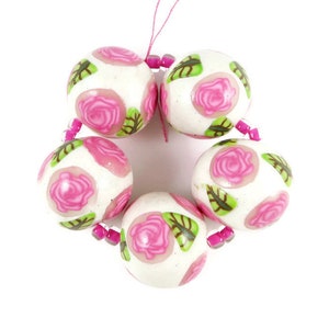 May include: A strand of white beads with pink roses and green leaves. The beads are round and have a glossy finish.