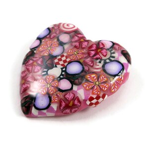 May include: A pink heart-shaped charm with a colorful floral pattern. The charm is made of polymer clay and features a variety of flowers, swirls, and polka dots in shades of pink, red, white, and purple.