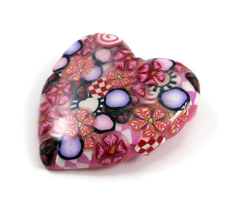 May include: A pink heart-shaped charm with a colorful floral pattern. The charm is made of polymer clay and features a variety of flowers, swirls, and polka dots in shades of pink, red, white, and purple.