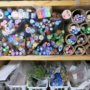 May include: A shelf filled with colorful polymer clay canes, organized in clear containers and cardboard tubes. The canes are arranged in various patterns and designs, including swirls, dots, and stripes.