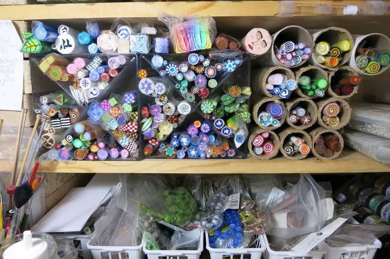 May include: A shelf filled with colorful polymer clay canes, organized in clear containers and cardboard tubes. The canes are arranged in various patterns and designs, including swirls, dots, and stripes.