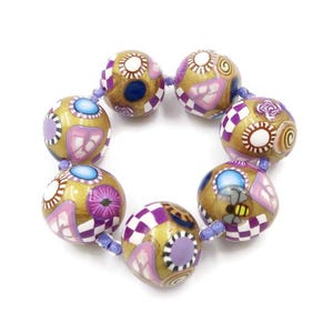 May include: A beaded bracelet featuring gold-colored, round beads with intricate designs in purple, white, and blue. The beads are connected by a light purple string, creating a circular shape. The designs include geometric patterns and floral motifs.