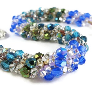 May include: A beaded bracelet featuring a mix of blue, green, and clear beads. The bracelet has a silver clasp and is made with various sizes and shapes of beads, creating a textured look. The beads have a shiny, iridescent finish.