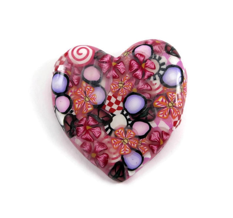 May include: A pink heart-shaped pendant with a colorful floral pattern. The pattern includes pink, red, white, and purple flowers, as well as swirls and polka dots.