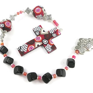 Puede incluir: Una cruz con estampado rojo y negro con un dije de pez y cuentas cúbicas negras en una cadena de cuentas blancas y rosadas.