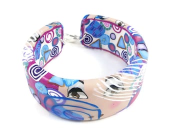 Op de afbeelding: Een brede, kleurrijke armband gemaakt van polymer clay. De armband heeft een roze, wit en blauw geometrisch patroon met swirls, cirkels en abstracte vormen. De armband is open en heeft een zilveren sluiting.