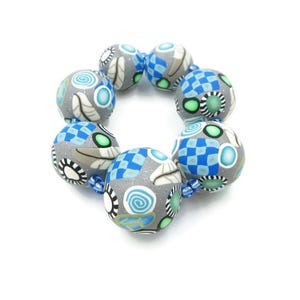 May include: A handmade bracelet featuring large, round beads in shades of gray, blue, and white. The beads have intricate patterns including swirls, checks, and floral designs. The bracelet is strung on a blue cord and is set against a white background.