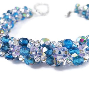 May include: A close-up of a beaded bracelet featuring a mix of clear, blue and purple beads. The bracelet is crafted with small seed beads and larger faceted beads.