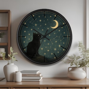 Puede incluir: Un reloj de pared redondo con marco negro. La esfera del reloj presenta una silueta de gato negro mirando una luna creciente dorada y estrellas sobre un fondo verde azulado oscuro. Las manecillas del reloj son negras.