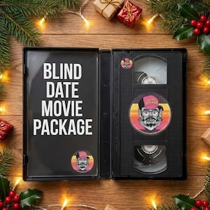 May include: An open black VHS case with the text "BLIND DATE MOVIE PACKAGE" on the left side. The right side contains a VHS tape and a circular sticker with a bearded man wearing glasses and a hat. The background is a wooden surface with Christmas decorations.