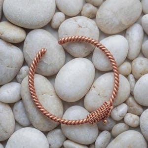 Op de afbeelding: Een koperkleurige metalen manchetarmband met een gedraaid, touwachtig ontwerp over de gehele band. De armband heeft een open stijl en bevat een decoratief gedeelte van strak gewikkeld draad nabij een van de uiteinden van de armband.