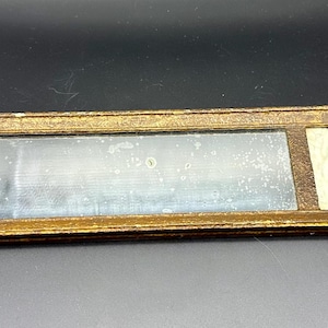 May include: A long, narrow, gold-toned picture frame with a mirror on the left side and a small painting on the right. The painting depicts a woman in a white dress with a blue sash, holding a small dog.